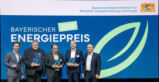Bayerns Wirtschaftsminister Hubert Aiwanger (rechts) und Moderator Sebastian Schaffstein (links) übergeben den Bayerischen Energiepreis 2026 für das Projekt „einfach.anders.machen.RESI“ an die Preisträger Alpha IC GmbH und Hölzlein Ingenieure GmbH: (v.l.n.r.) Thomas Kohmann, Prokurist der Alpha IC GmbH, Christian Hölzlein, geschäftsführender Gesellschafter Hölzlein Ingenieure, Jan Hesse, Partner Alpha IC GmbH.